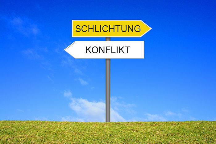Wegweiser auf der Wiese vor blauem Himmel. Schild nach rechts zeigt Schlichtung, Schild nach links zeigt Konflickt Wegweiser auf der Wiese vor blauem Himmel. Schild nach rechts zeigt Schlichtung, Schild nach links zeigt Konflickt